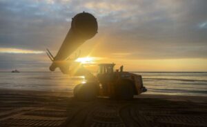 Bulldozer-like construction machine on a sandy beach at sunset, lifting a large cylindrical pipe toward the sky over calm sea water.