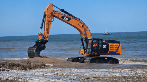 Orange CAT excavator on a sandy beach digging into the shore with the sea in the background.